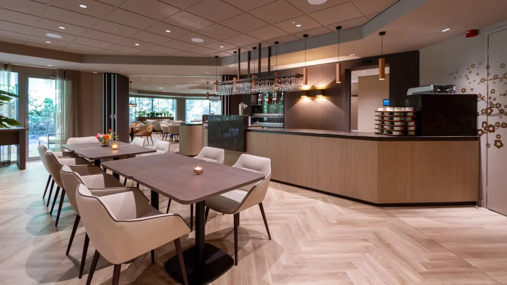 Rechthoekige tafels met beige stoelen voor lange houten bar in Crematorium Venlo; grote ramen en visgraatvloer.