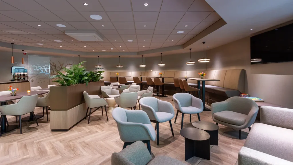 oungehoek in Crematorium Venlo met pastelblauwe fauteuils, lage tafels, televisiescherm en lange wandbank.