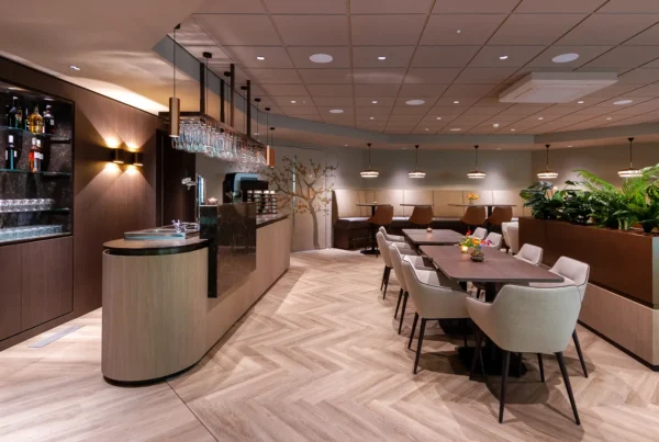Bar met glazenrek en koffiekamers in warme houttinten; centraal een groene plantenbak als rustige afscheiding.