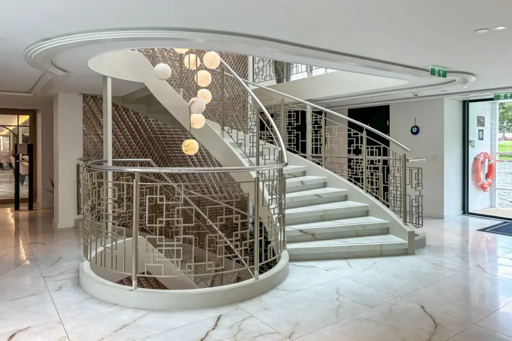 Centrale trap met gebogen metalen balustrades, sculpturale verlichting en warme wandaccenten creëren een herkenbare aankomstbeleving in de lobby van Excellence Crown.
